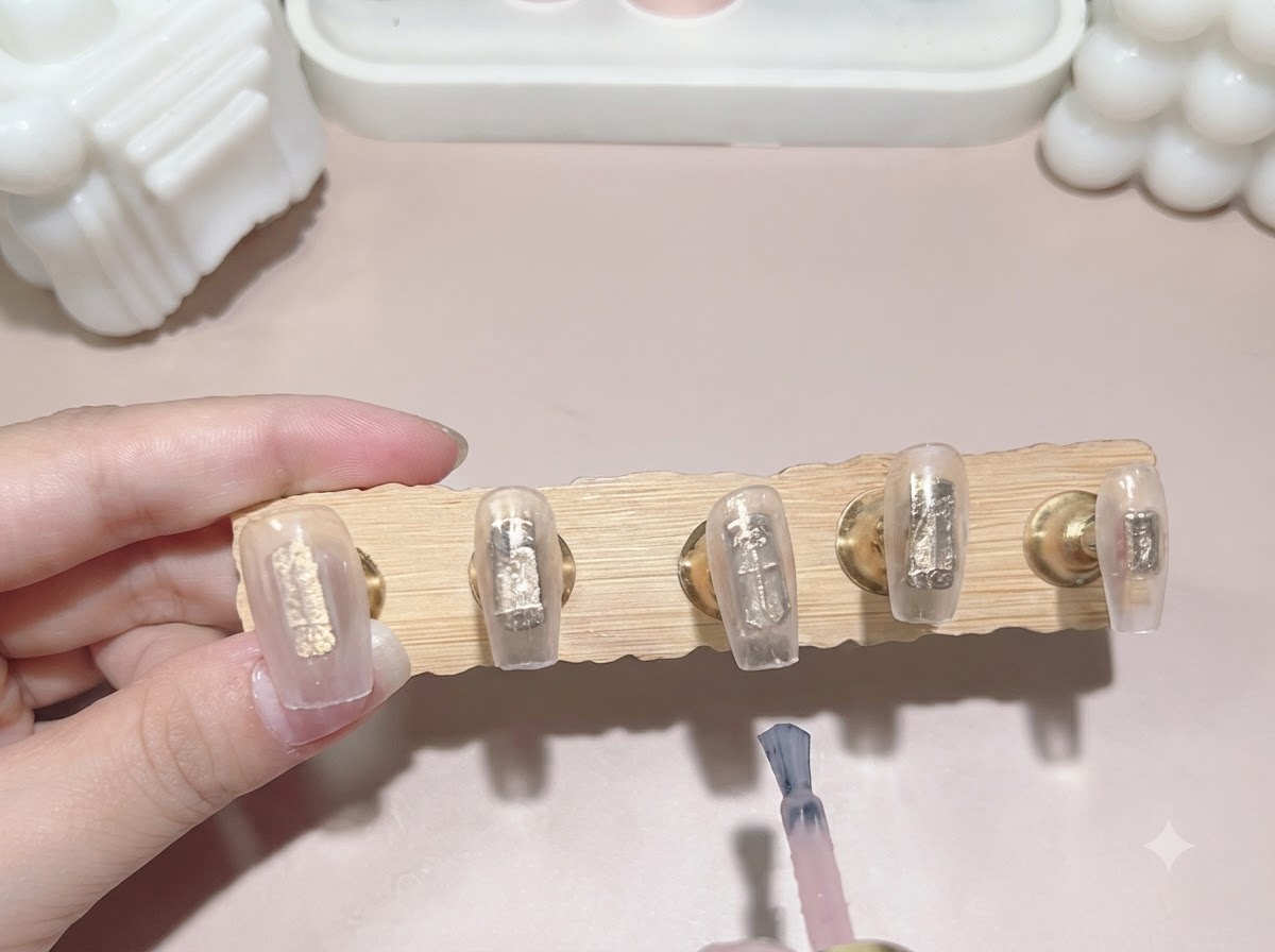 Ein Gel-Polish-Pinsel trägt eine zweite Schicht milchig-transparenter rosa Farbe auf Nagelspitzen auf, die an einem Holz-Display-Ständer befestigt sind.