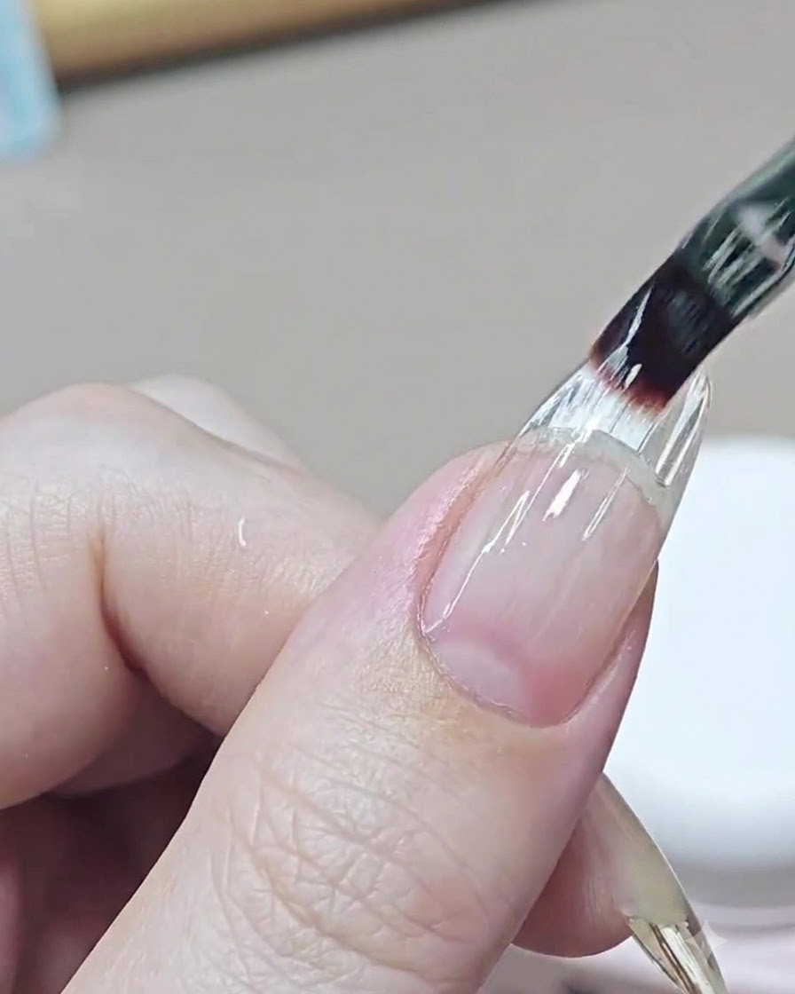 Ein feiner Nagelpinsel trägt eine klare Ausgleichsschicht Builder Gel über den Übergangsbereich eines verlängerten Nagels auf.