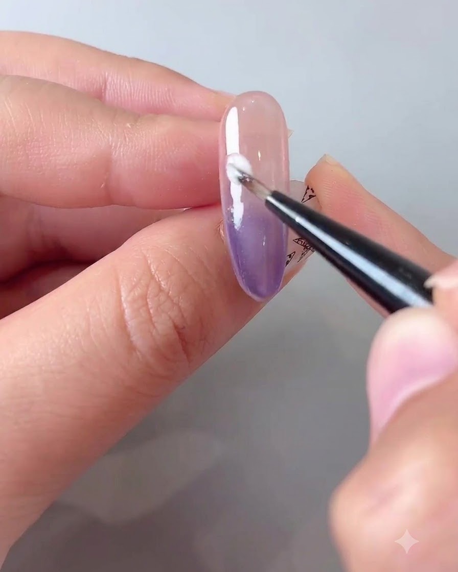 Ein feiner Pinsel, der eine kleine Menge milchweißes Gel diagonal über einen lila Farbverlaufsnagel aufträgt.