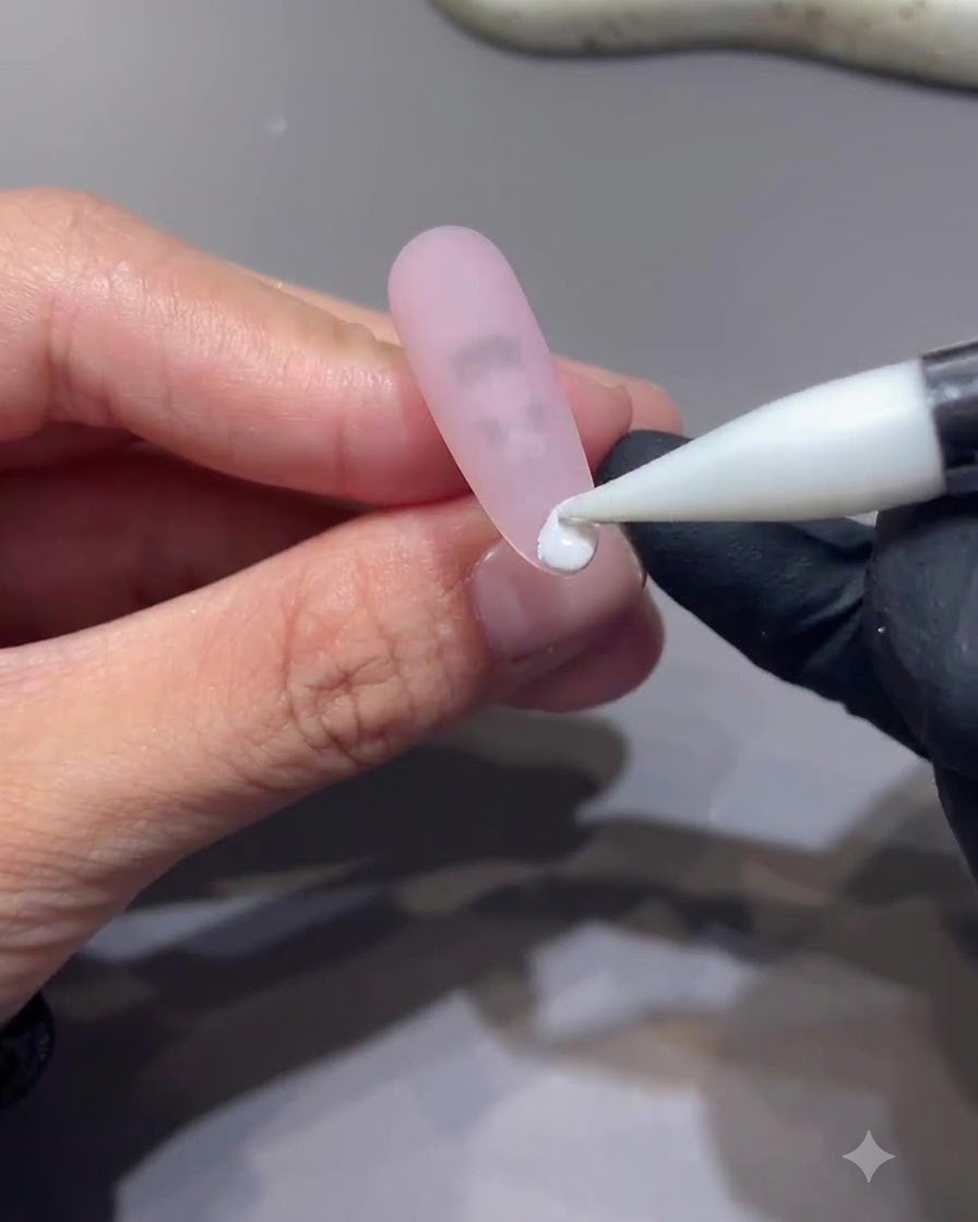 Ein Feinarbeitswerkzeug platziert einen kleinen Tropfen weißes Gel auf die Spitze eines matten rosa Nagels.