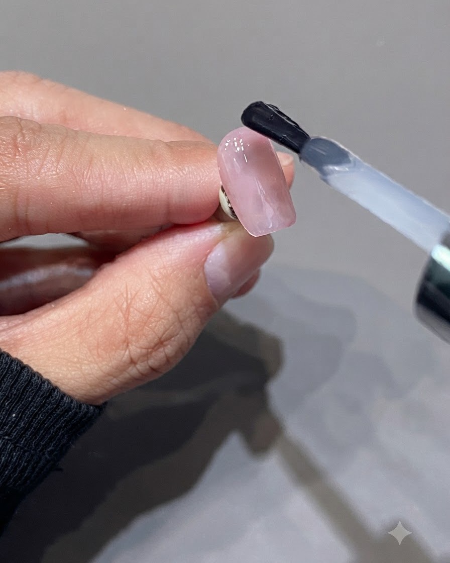 Ein Pinsel trägt einen transluzenten, milchig-rosafarbenen Top Coat auf eine glänzende rosa Nageloberfläche auf.