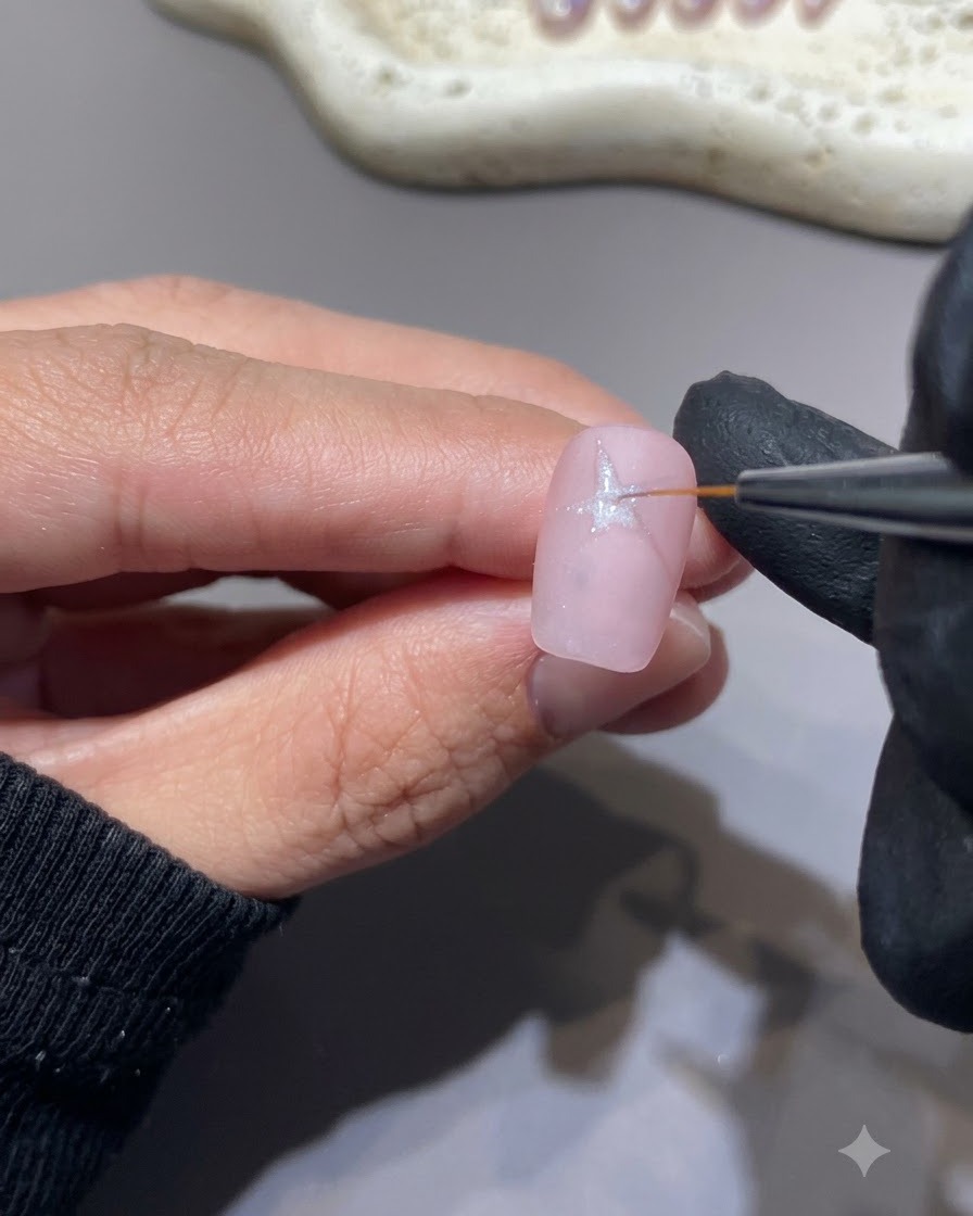 Ein feiner Liner-Pinsel zeichnet eine weiße Sternform auf eine rosa Nageloberfläche.