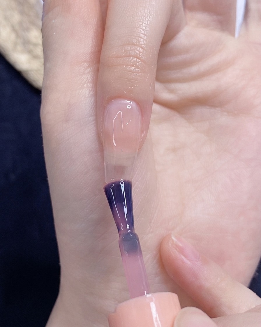 Ein Pinsel trägt eis-transluzenten Rosa-Gelack gleichmäßig auf einen unlackierten Fingernagel auf.