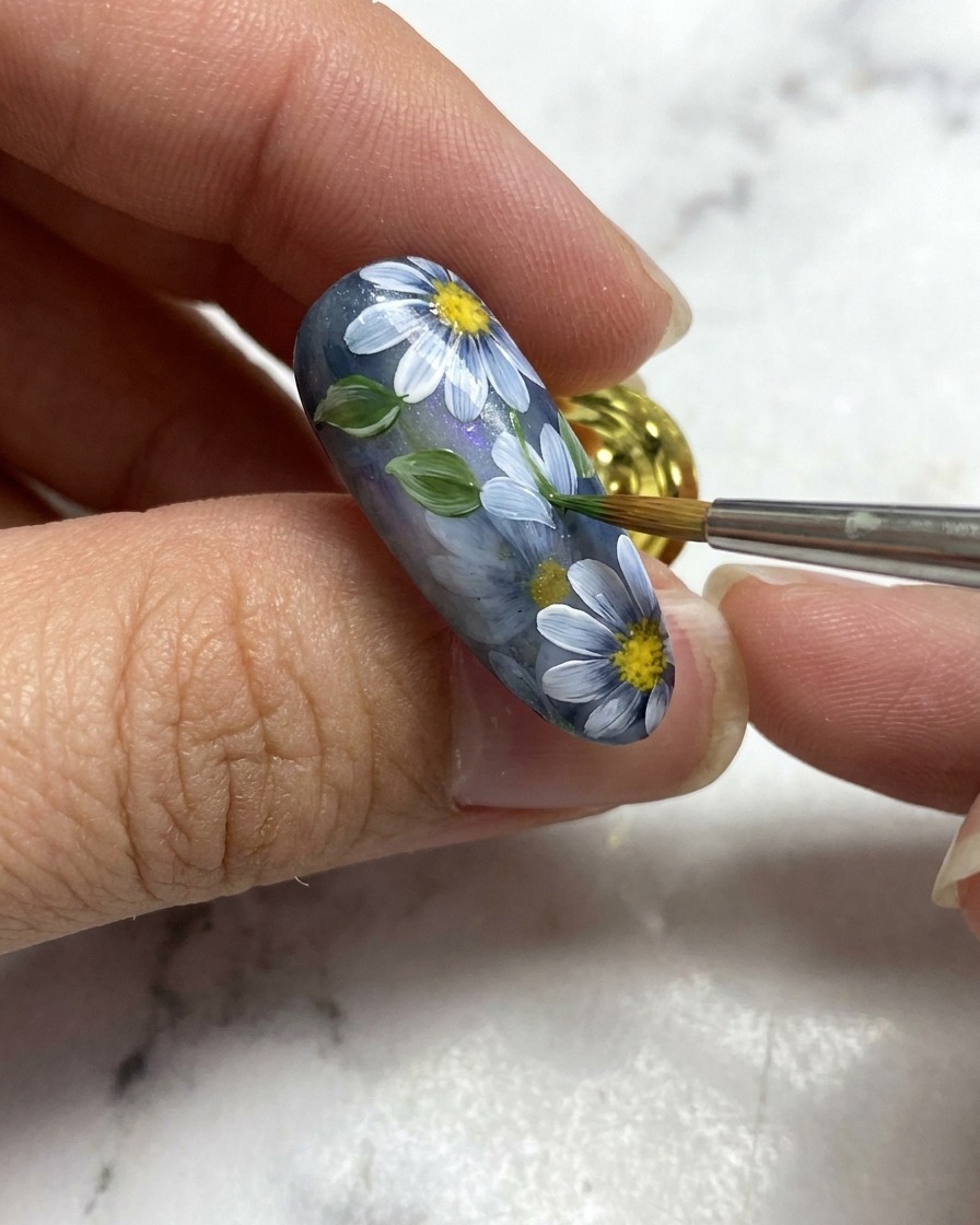 Ein Nail-Art-Pinsel fügt ein kleines grünes Blatt neben weißen Blumendetails auf einer grauen Nagelbasis hinzu.