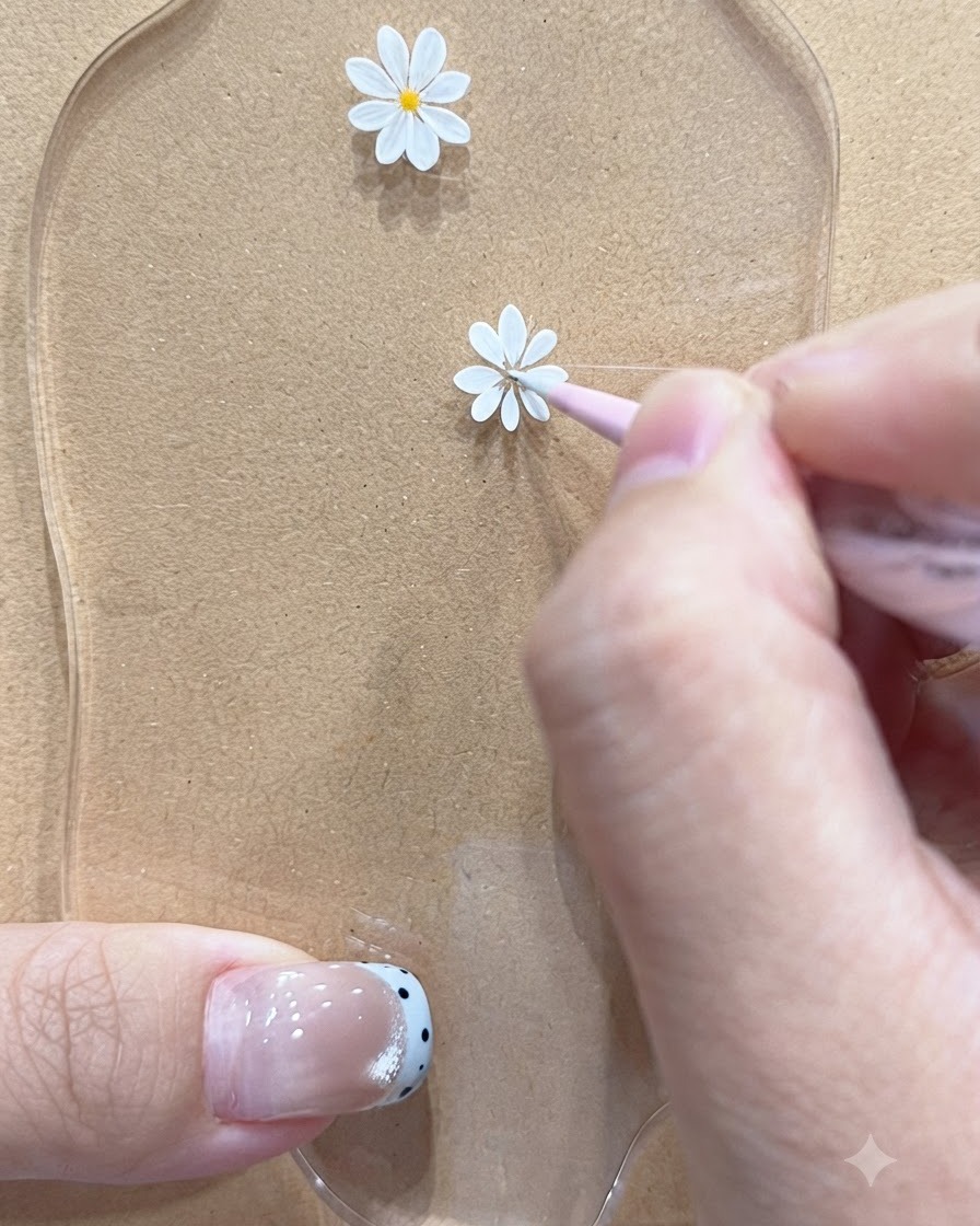 Ein Fine-Liner-Pinsel zeichnet zarte Blütenblätter für ein kleineres Gänseblümchen-Motiv auf einer Palette.