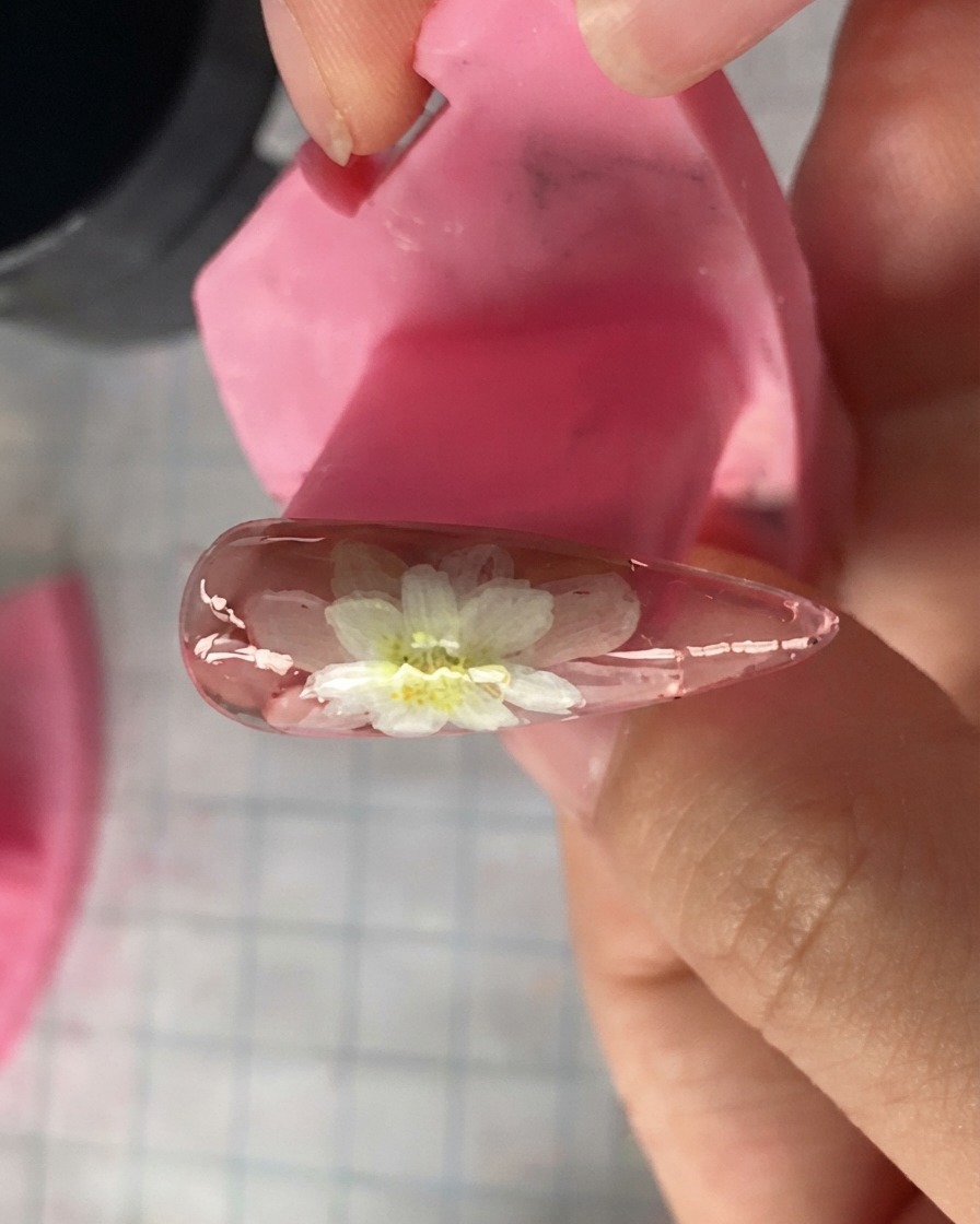 Eine Nahaufnahme eines rosa Nagelmodels zeigt geschichtete weiße Gänseblümchen-Blütenblätter, die oben aufgemalt sind.