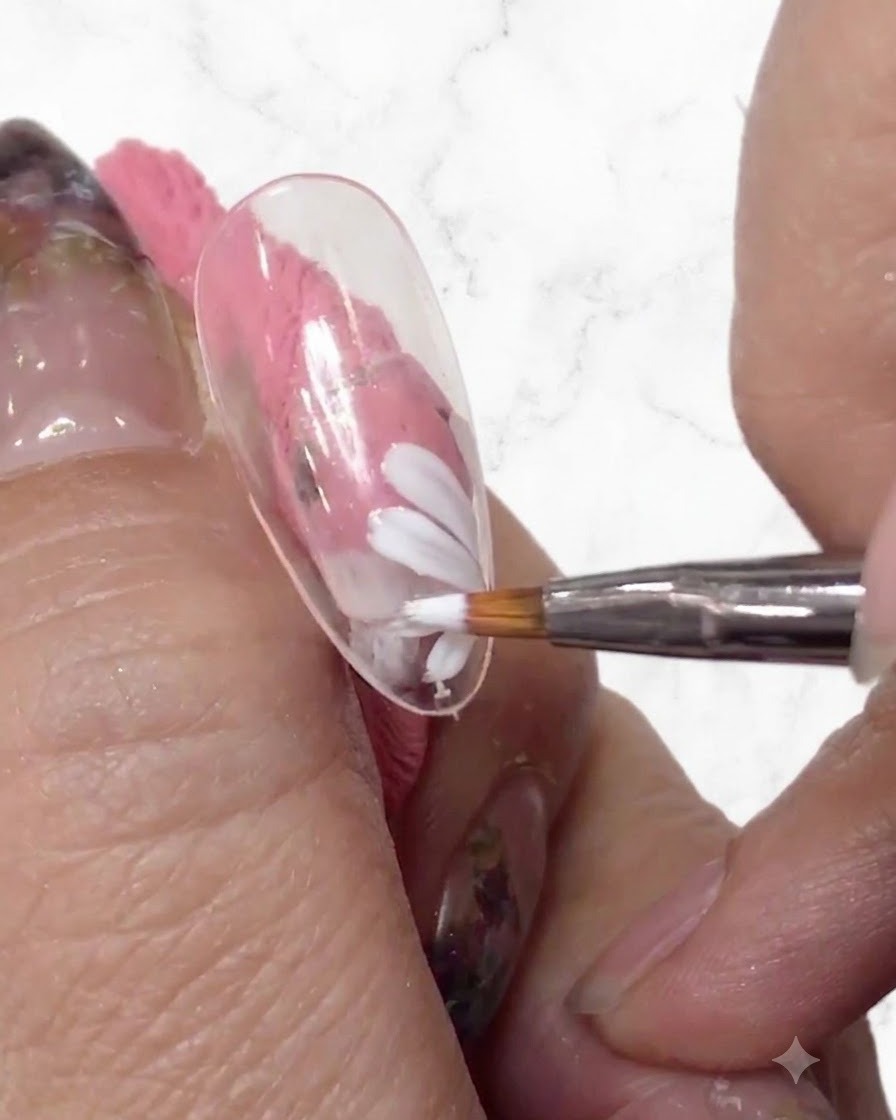 Eine Nahaufnahme eines Nageldesigners, der mit einem feinen Pinsel weitere weiße Blütenblätter zum Gänseblümchen-Design auf einem klaren Nagel-Tip hinzufügt.