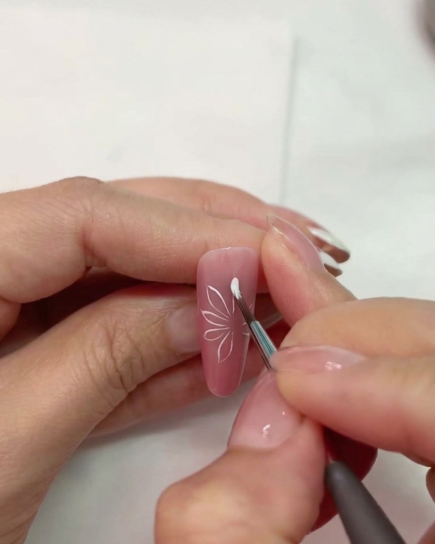 Nahaufnahme eines Nagelpinsels beim Zeichnen weißer Blütenblattformen auf einem pinken matten Nagel.