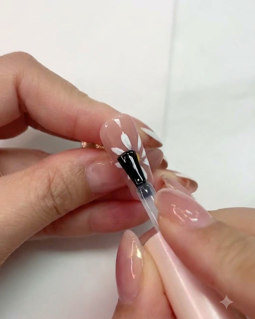 Ein Pinsel trägt matten Topcoat über ein ausgehärtetes weißes Gänseblümchendesign auf einem pinken Nagel auf.