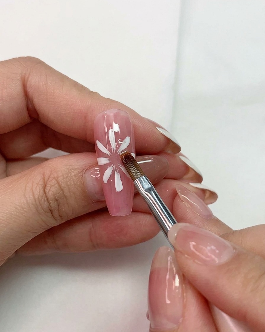 Eine Hand hält einen Mehrzweckpinsel zum Formen weißer Blütenblätter über einer pinken Basis auf einem Fingernagel.