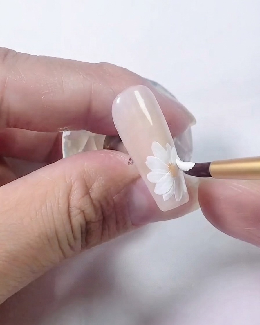 Ein Nagelpinsel, der eine zweite, überlappende Schicht weißer Blütenblätter auf das Gänseblümchen aufträgt.