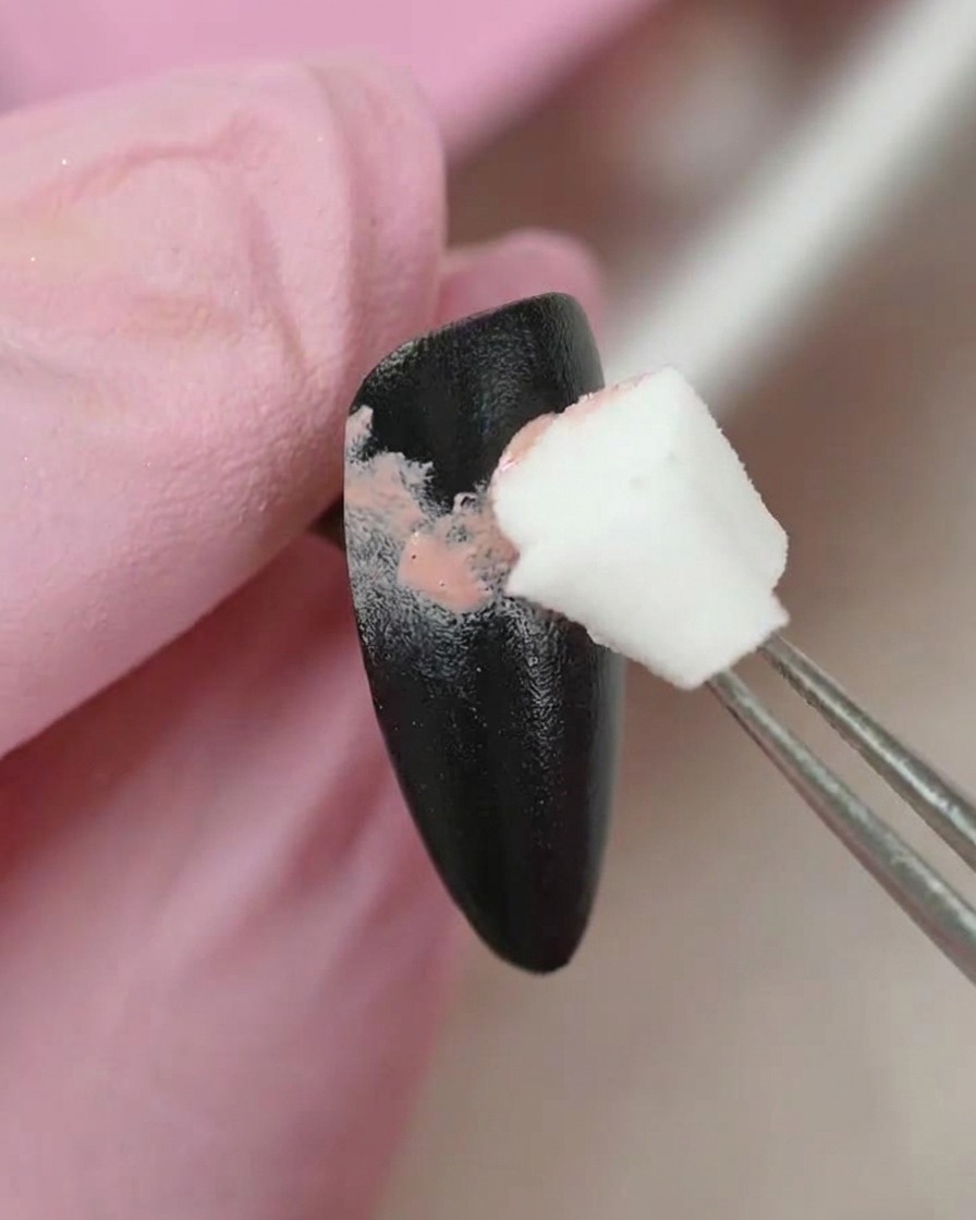Eine Pinzette hält einen kleinen Schwamm-Applikator und tupft sanft pinke Gel-Farbe auf eine schwarz lackierte Nageloberfläche.