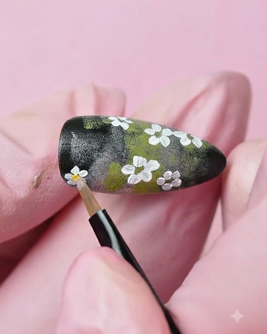 Ein feiner Detailpinsel trägt einen Tropfen gelbes Gel auf die Mitte einer gemalten weißen Blume auf dem Nagel auf.
