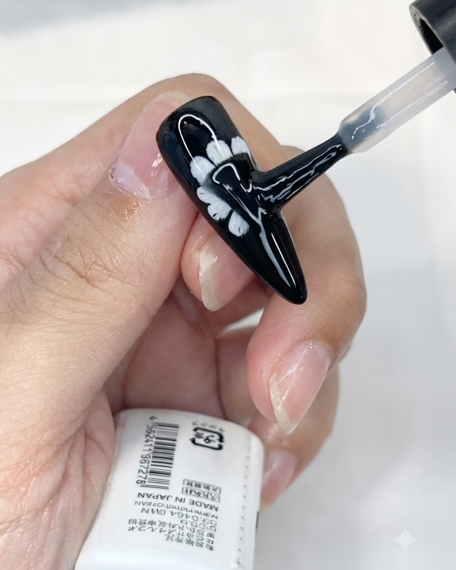 Ein Pinsel trägt eine Schicht matten Topcoat auf einen schwarzen Nagel mit weißem Gänseblümchen-Design auf.