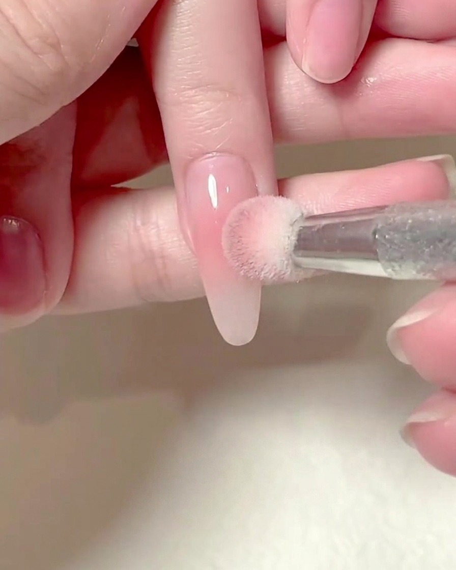Ein quadratischer Schwamm, gehalten von einer Hand, die sanft rosafarbenes Blush-Gel auf einen verlängerten Fingernagel tupft.