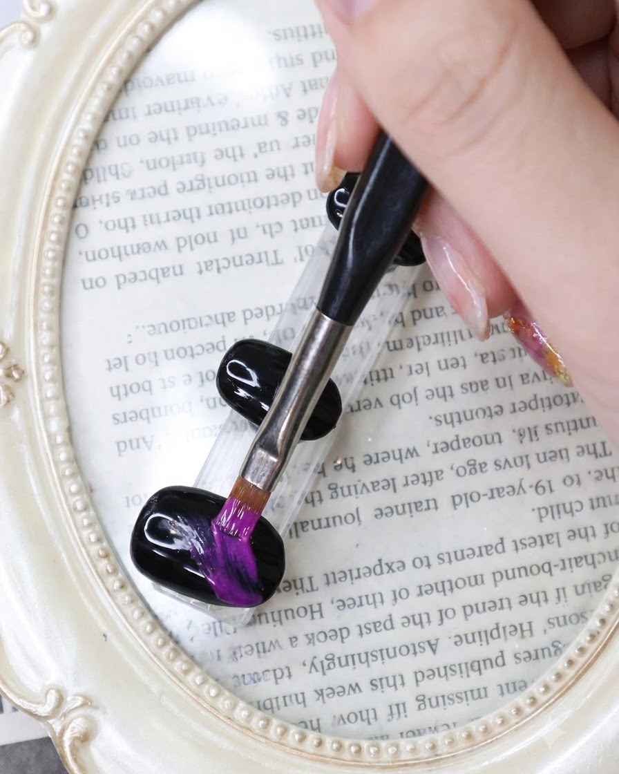 Ein feiner Liner-Pinsel zieht leuchtend neonviolette Pinselstriche über einen schwarzen Gel-Nagel vor einem buchbezogenen Hintergrund.