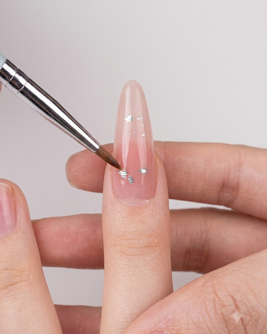 Ein feiner Nail-Art-Pinsel platziert kleine silberne irisierende Flocken auf einen nudefarbenen, glänzenden Fingernagel.