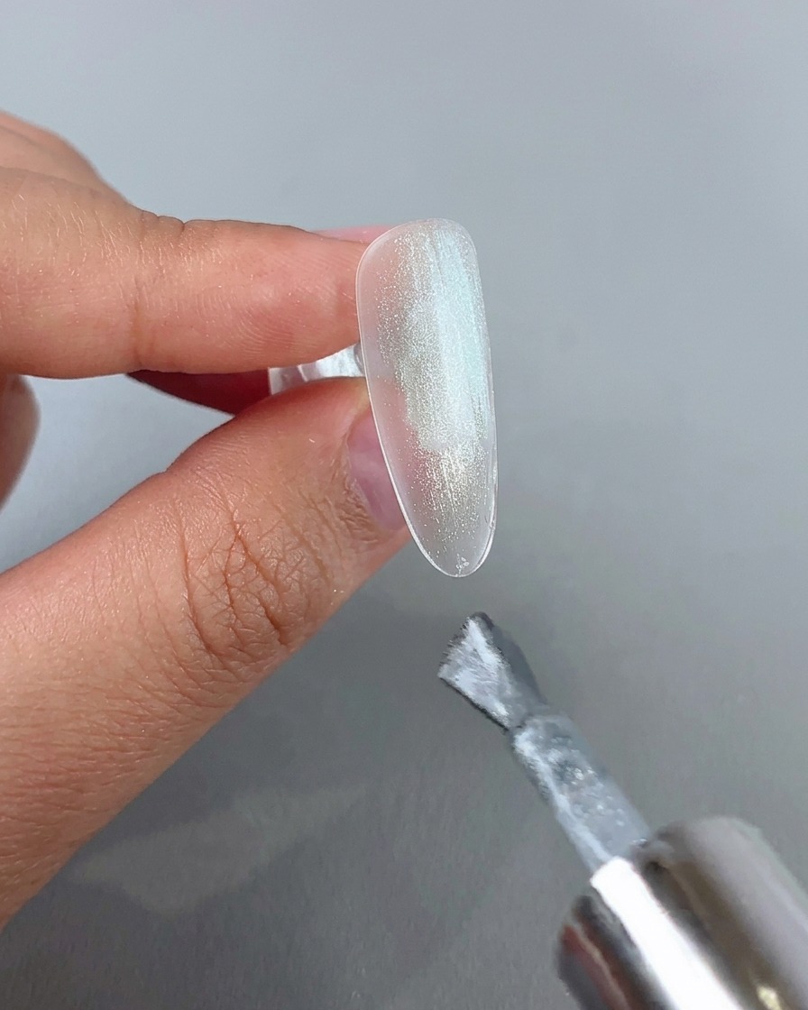 Ein Nagelpinsel trägt eine Schicht glitzerndes Silber-Cat-Eye-Gel auf eine klare Nagelspitze auf.