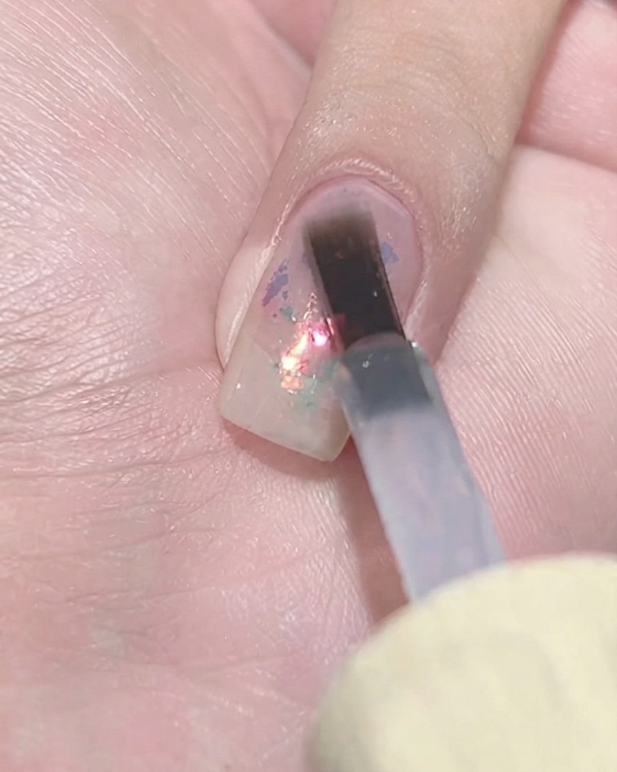 Ein flacher Gel-Pinsel tupft eine Schicht transparenten violetten Lack über ein strukturiertes Flake-Cluster auf einem Fingernagel.