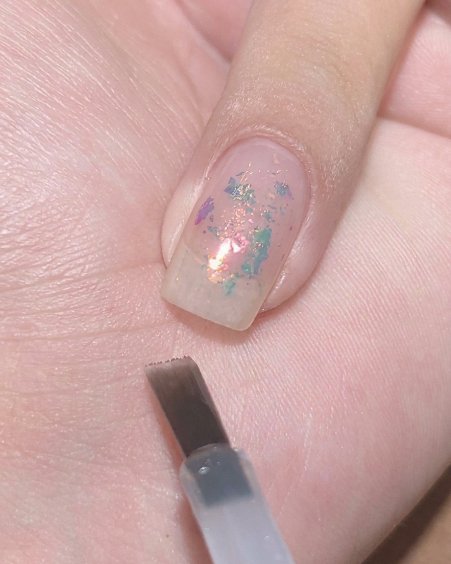 Ein flacher Gel-Pinsel schwebt nahe an einem klaren Nagel mit eingebetteten Flakes, bereit zum Auftupfen von transparentem violetten Lack.