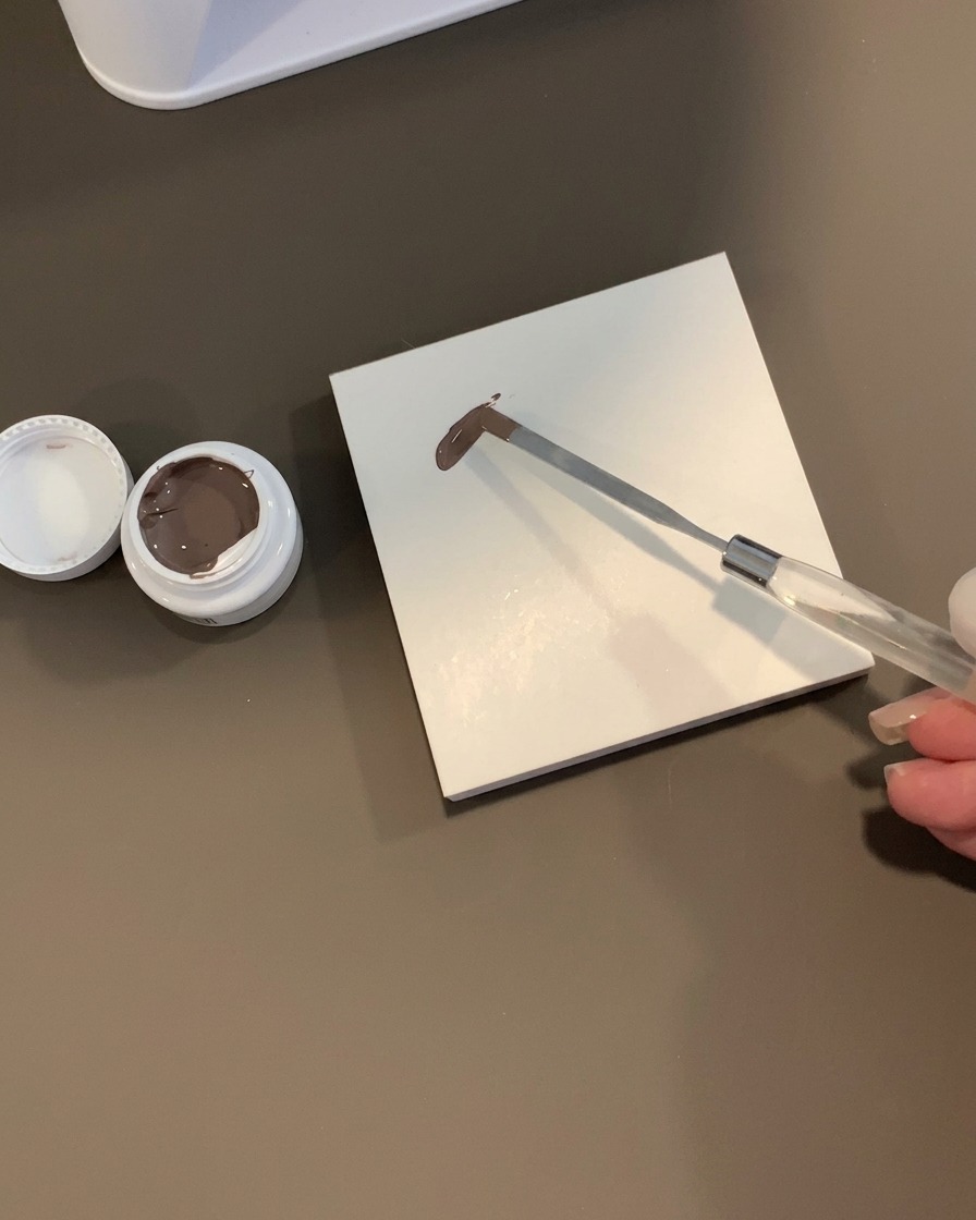 Ein Metallsputel hält einen Klecks braungraues Gel auf einer weißen quadratischen Fliesenpalette neben einem offenen Tiegel.