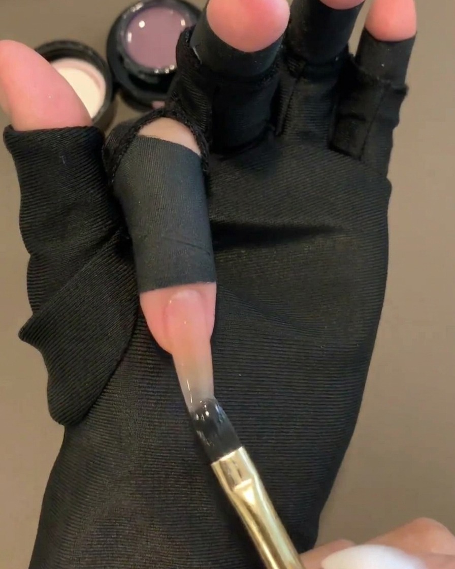 Ein flacher Gelpinsel trägt ein transparentes violettes Farbgel auf die obere Hälfte eines Nude-Basis-Nagels auf.