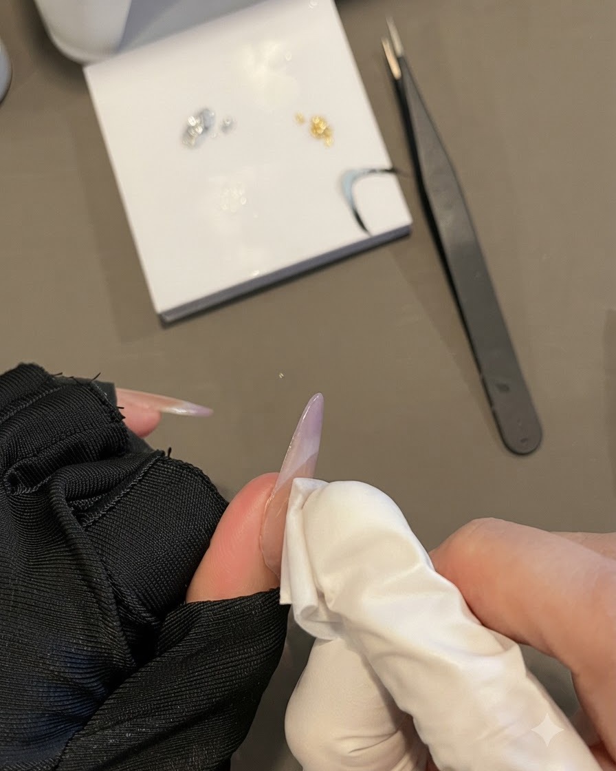 Eine behandschuhte Hand wischt eine Nageloberfläche mit einem fusselfreien Tuch ab, während sie einen langen ovalen Nagel hält.