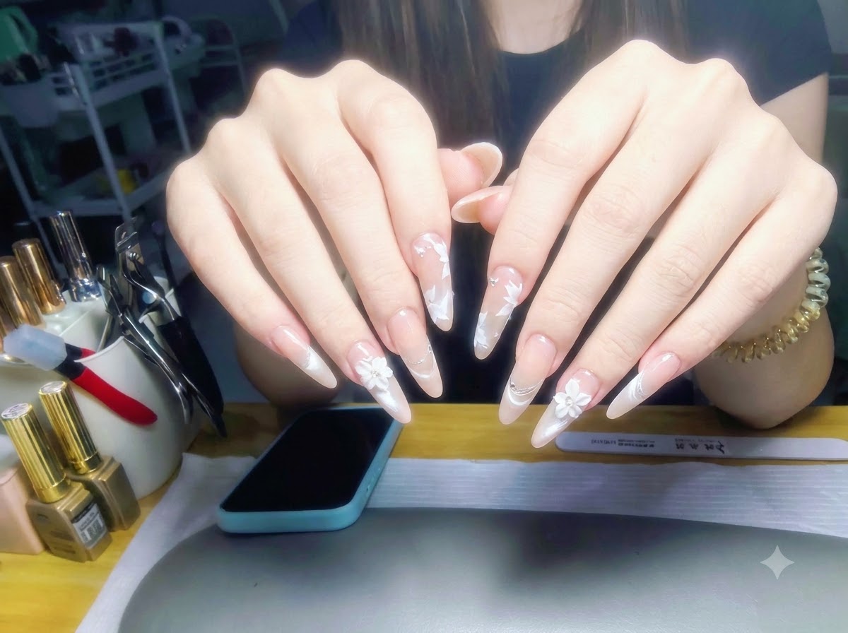 Beide Hände vor einem Arbeitsplatzhintergrund erhoben, zeigen ein vollständiges Set glänzender Nude-Perlmutt-Gradienten-Nägel mit weißer Blumenkunst.