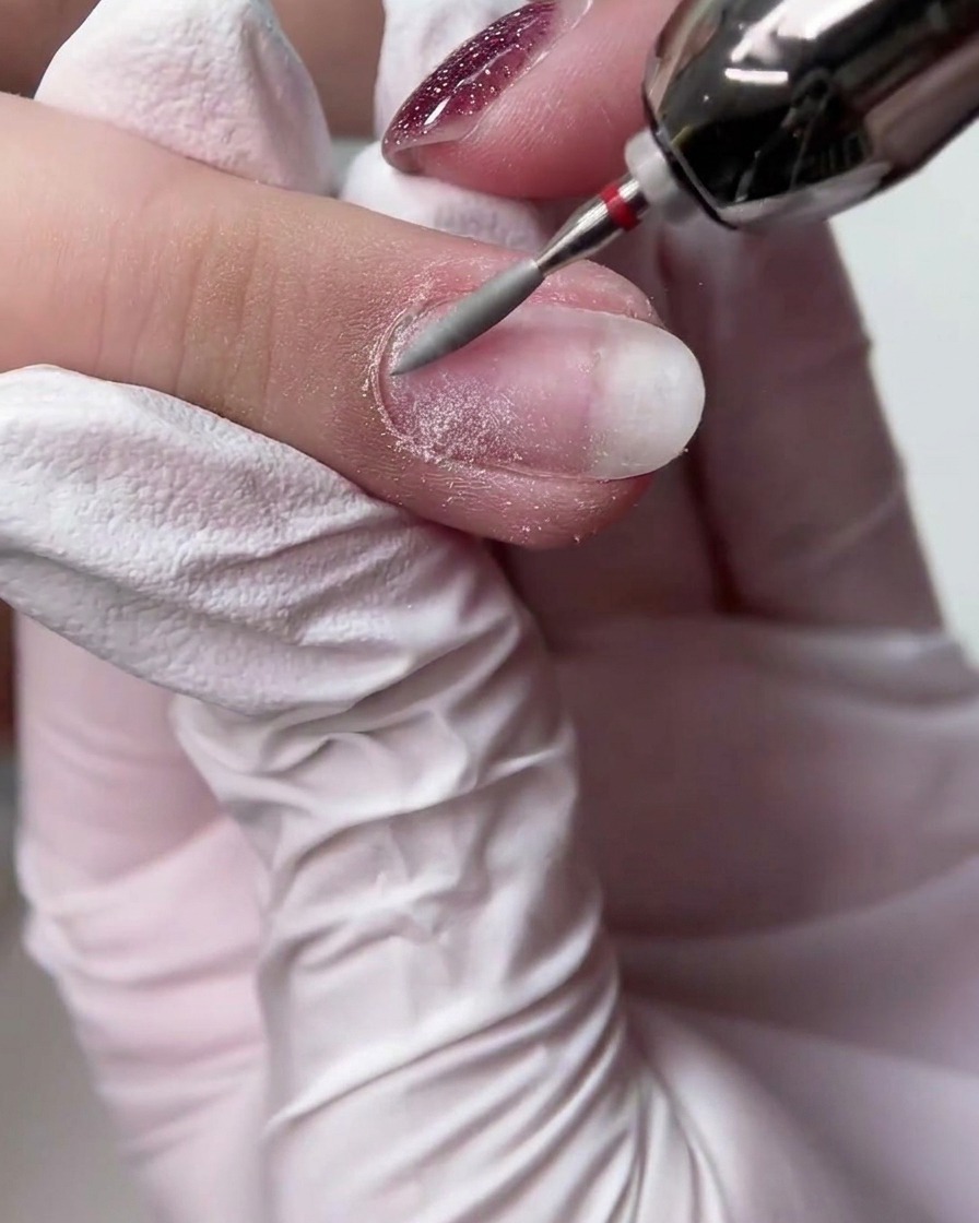 Eine behandschuhte Hand verwendet einen Fräser mit langem konischen Bit, um die Oberfläche eines natürlichen Fingernagels zu glätten.