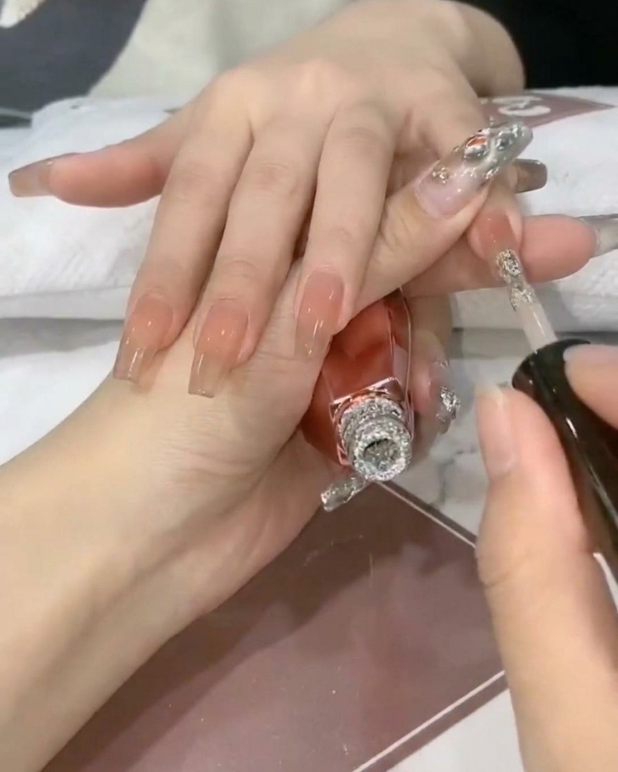 Ein Nagelpinsel gibt funkelndes Silberglitzer-Gel auf die Spitze eines transparenten Nude-Nagels.