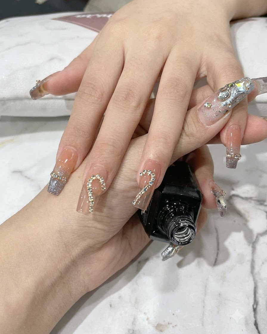 Eine Pinzette setzt kleine, funkelnde Strasssteine in eine Metallkettenumrandung auf einem Fingernagel.