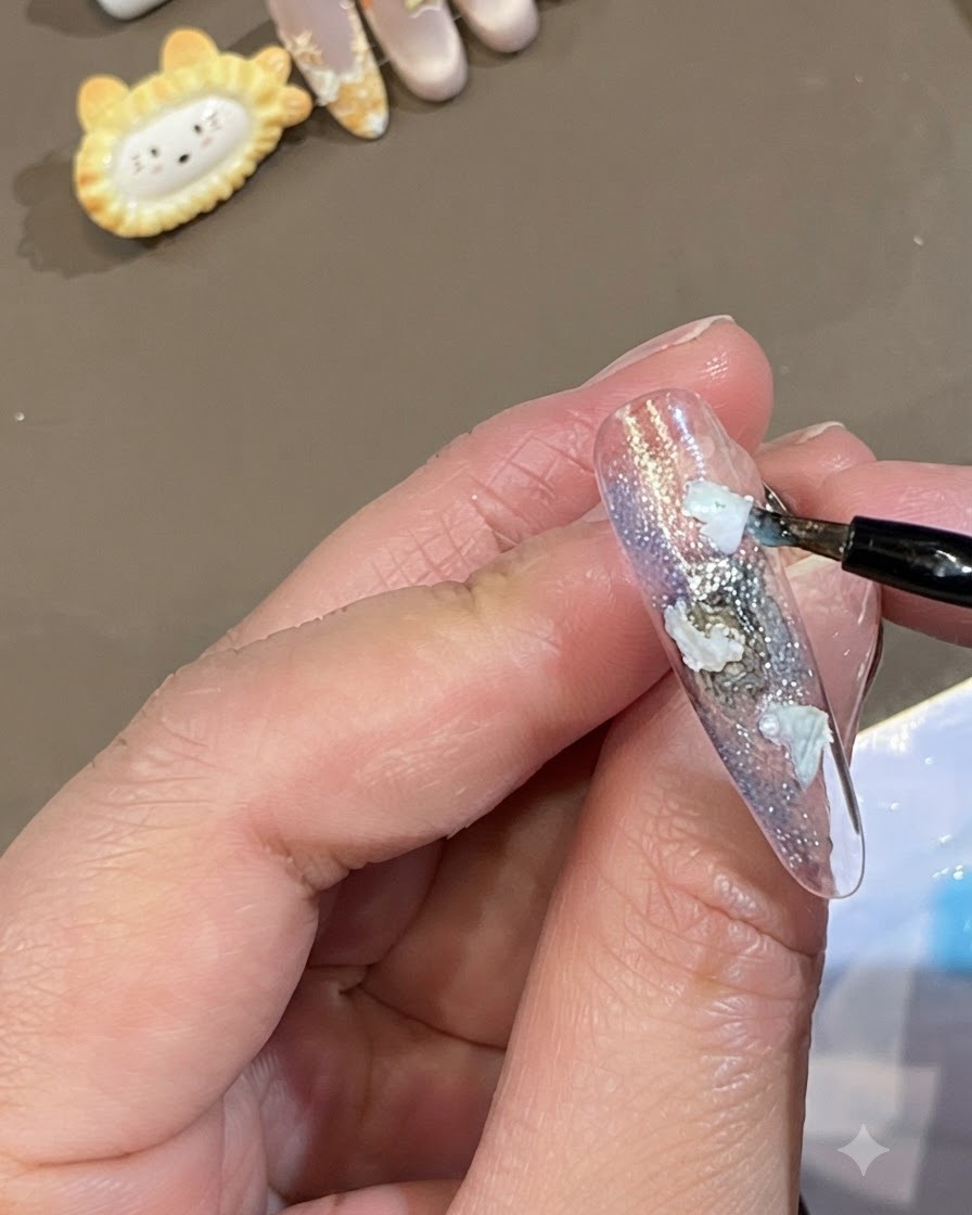 Ein feiner Pinsel setzt Punkte aus transluzentem blauem Gel um die Kanten weiße Muschelstücke auf einem Nagel.