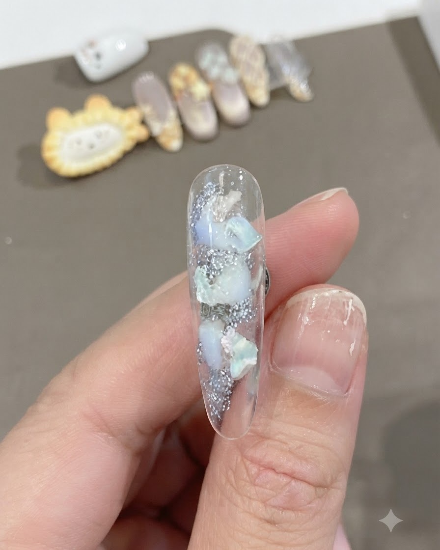 Eine Nahaufnahme eines klaren, langen Nagels mit eingebetteten Muschelstücken und schimmernder Textur mit einem weichen, weißen Farbverlauf.