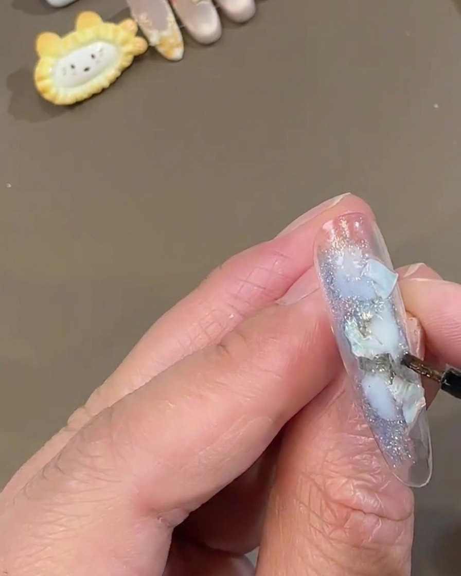 Ein feiner Detail-Nagelpinsel tupft weißen, funkelnden Feenstaub auf einen klaren Press-On-Nagel, der mit blauen Muschelstücken dekoriert ist.