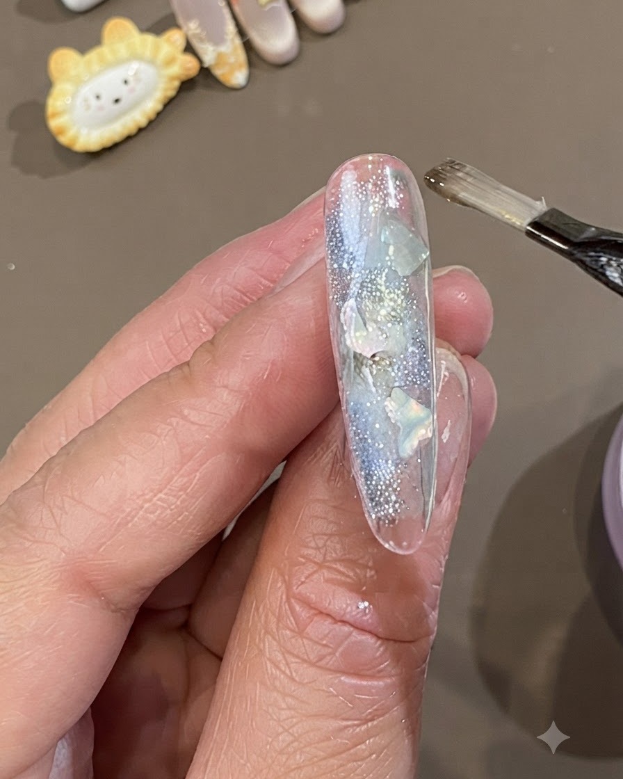 Ein flacher Gelpinsel trägt eine dicke Schicht klares Soft-Versiegelungsgel über einen mit Muschelstücken und Glitzer verzierten Nagel auf.