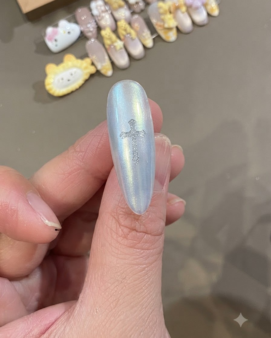 Eine Nahaufnahme eines perlmuttartigen weissen Nagels mit einem funkelnden silbernen Chrome-Heart-Kreuz-Sticker direkt in der Mitte.