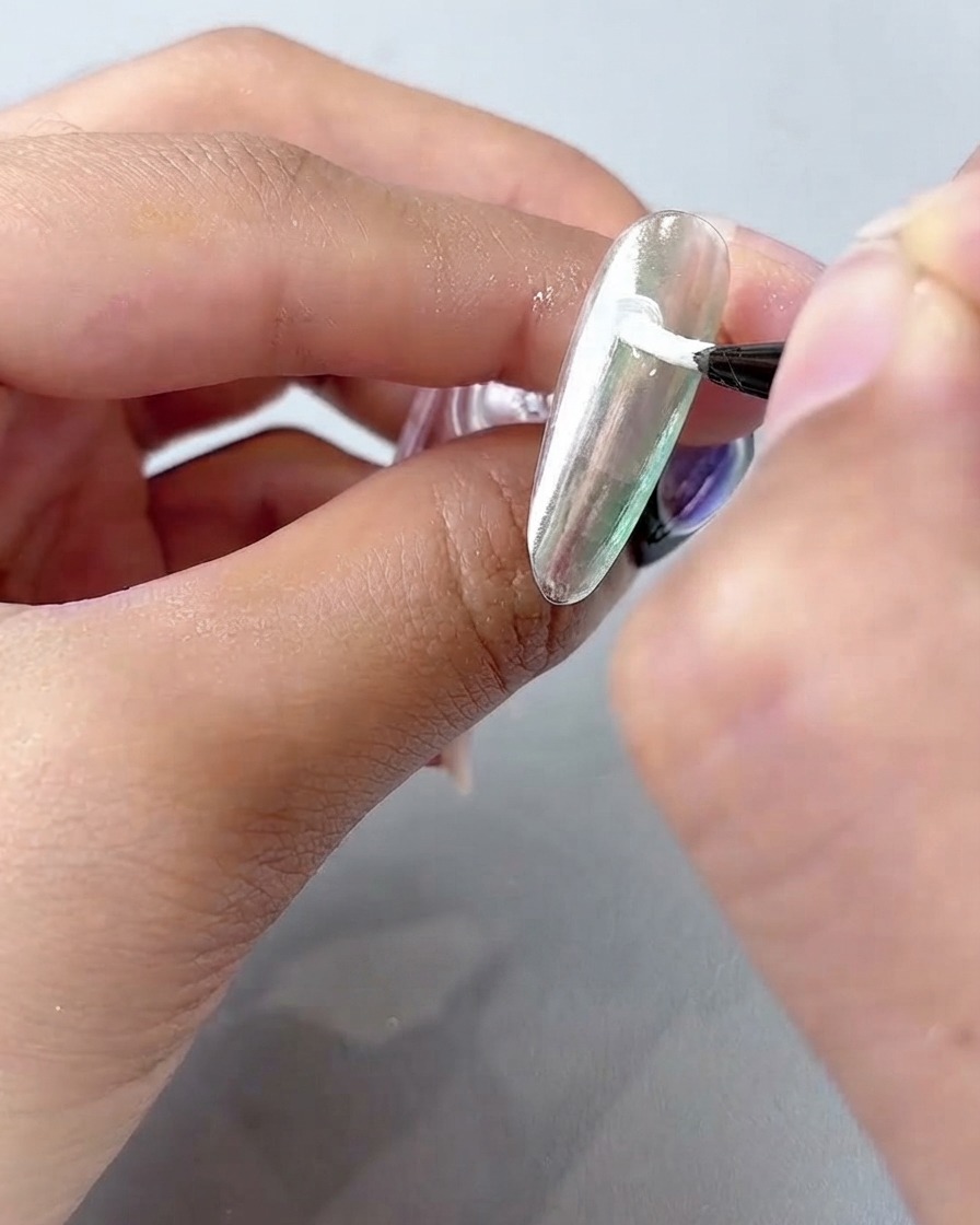 Ein feiner Nailart-Pinsel trägt weißes Malgel in gebogenen Blütenblättern auf eine silbrige Chrom-Nageloberfläche auf.
