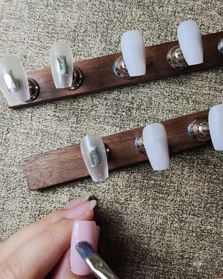 Ein flacher Gelpinsel trägt eine zweite Schicht zartrosa Nude-Gellack auf den Nagel auf.