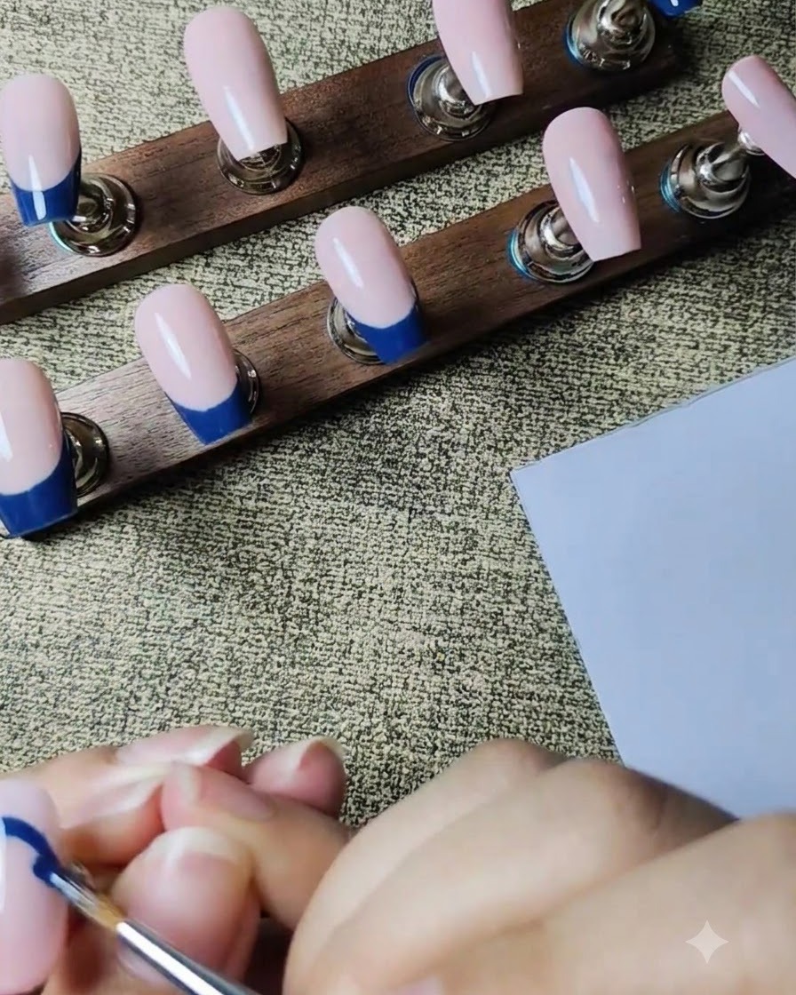 Ein feiner Liner-Pinsel zeichnet eine dunkelblaue French-Tip-Kontur auf einen nudefarbenen Press-On-Nagel.