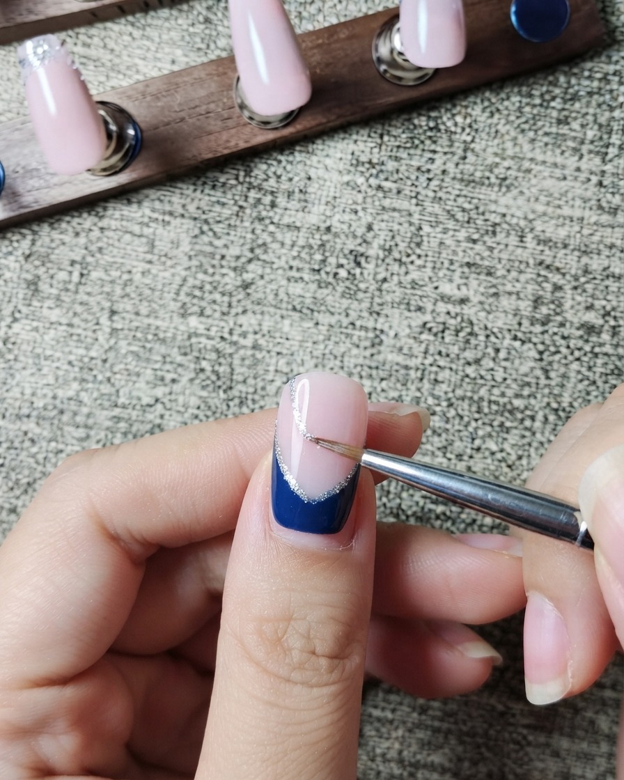 Ein Liner-Pinsel malt vorsichtig eine feine silberne Glitzerlinie entlang der inneren Kante eines dunkelblauen French Tips.