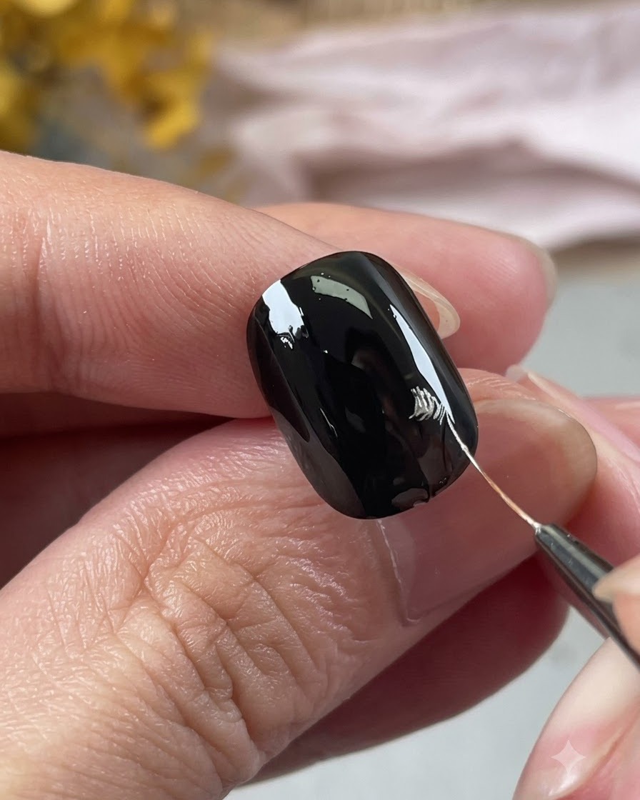 Eine Nahaufnahme einer Hand, die mit einem feinen Nail-Art-Pinsel silberne Blütenblattkonturen auf einen schwarzen Nagel zeichnet.