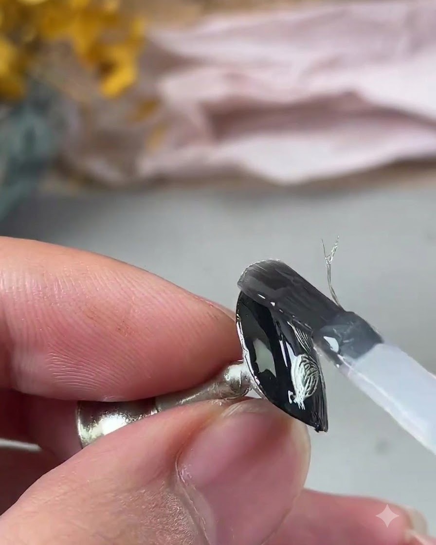 Ein Pinsel trägt eine klare, verstärkende Gelschicht über das texturierte Metallic-Nageldesign auf.