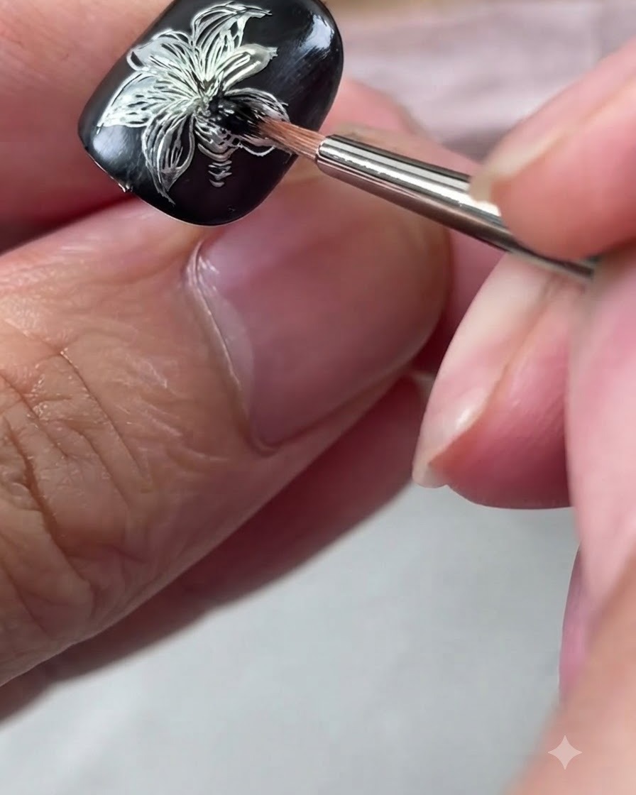 Ein Detailpinsel verblendet schwarze Gelpolitur im Zentrum eines silbernen Metallic-Lilien-Nageldesigns.
