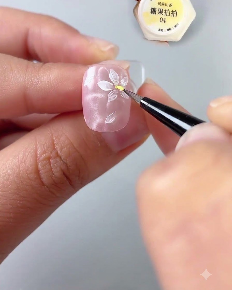 Nahaufnahme einer Hand, die einen Nagel hält und mit einem feinen Detailpinsel einen Punkt gelbes Gel in die Mitte einer gemalten weißen Blume auf einer glänzenden rosa Nagelbasis aufträgt.
