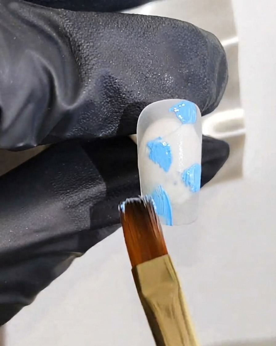 Eine behandschuhte Hand hält einen feinen Malpinsel und trägt blaue Gel-Punkte auf einen weißen Nagelgrund auf.