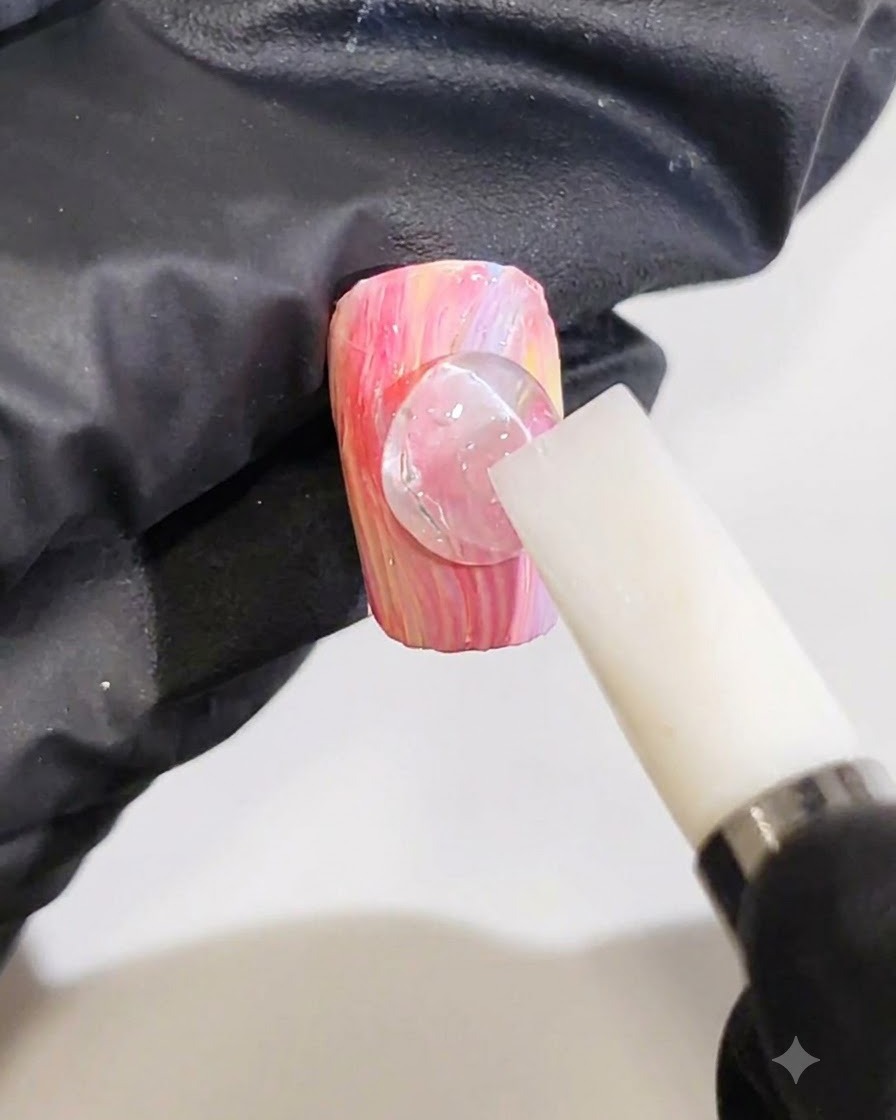 Eine behandschuhte Hand formt mit einem weißen Silikonwerkzeug einen klaren 3D-Geltropfen zu einer Wellenlinie auf einem pink-gelben Marmornagel.
