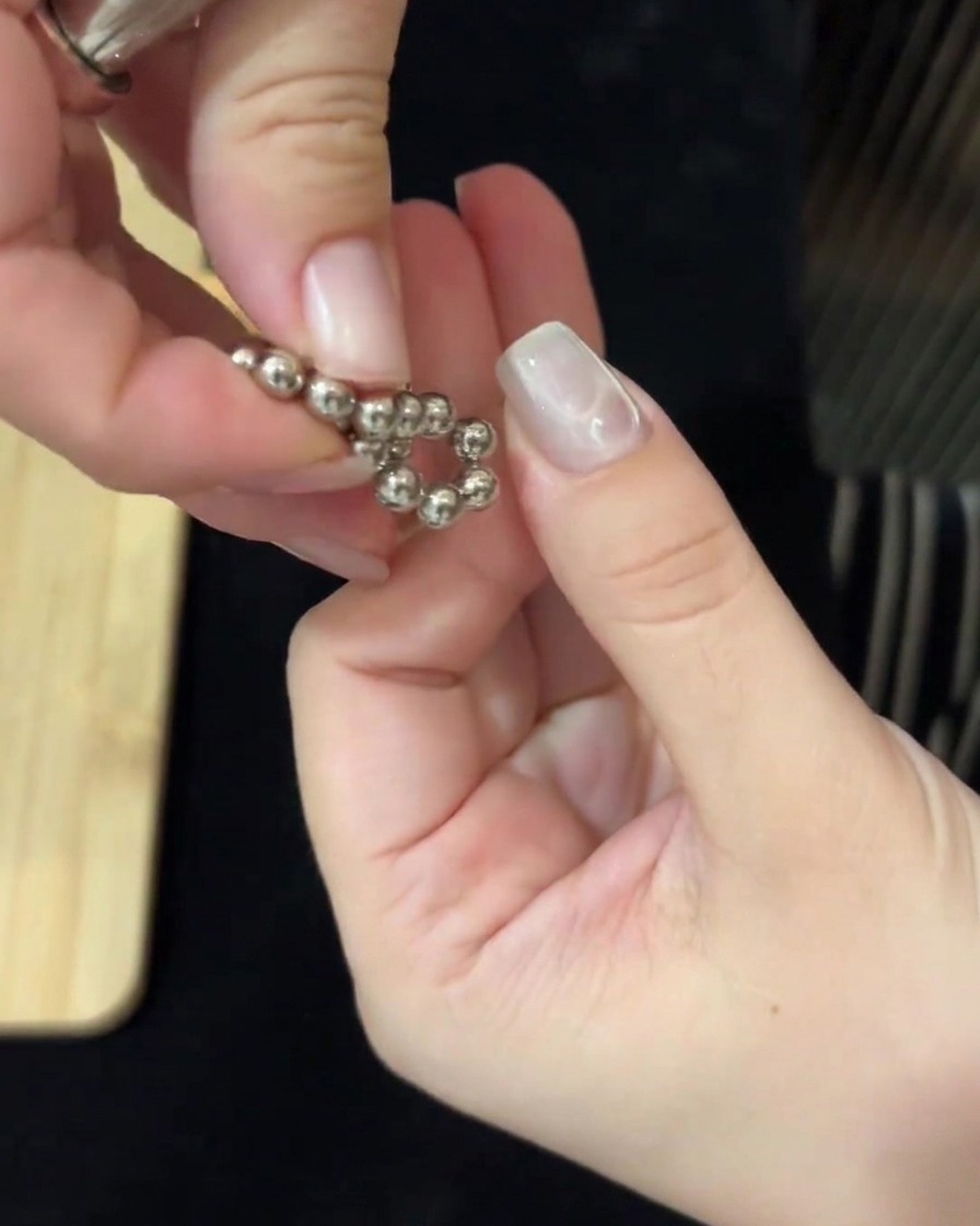 Hände nutzen magnetische Werkzeuge auf beiden Seiten, um metallische Perlen zu manipulieren und Wasserwellen auf dem Nagel zu erzeugen.