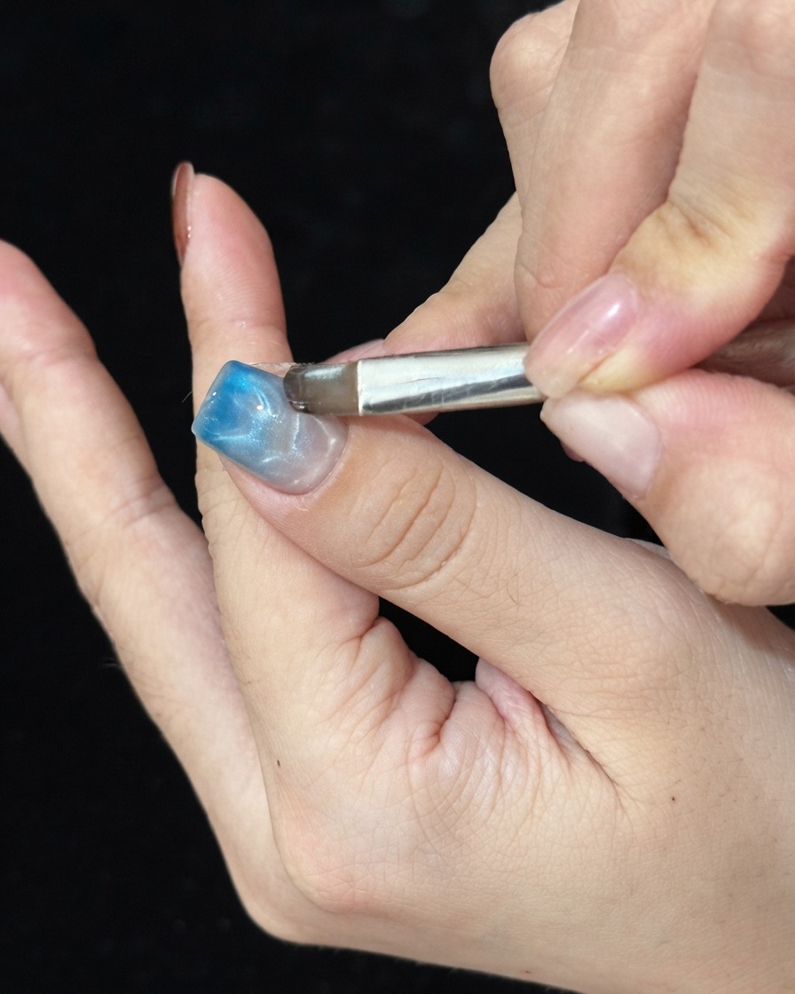 Ein Pinsel tupft blaues Glasgel auf die Spitze eines manikürten Nagels, um einen Ombré-Farbverlauf zu erzeugen.