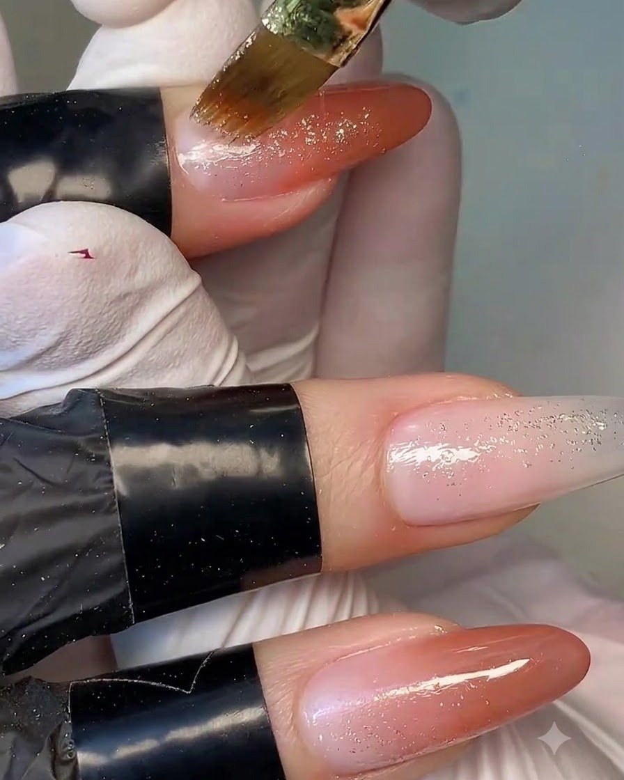 Ein feiner Pinsel trägt schimmernde Goldglitterpartikel auf die Nagelspitze und die Übergangszone des Farbverlaufs-Nagels auf.