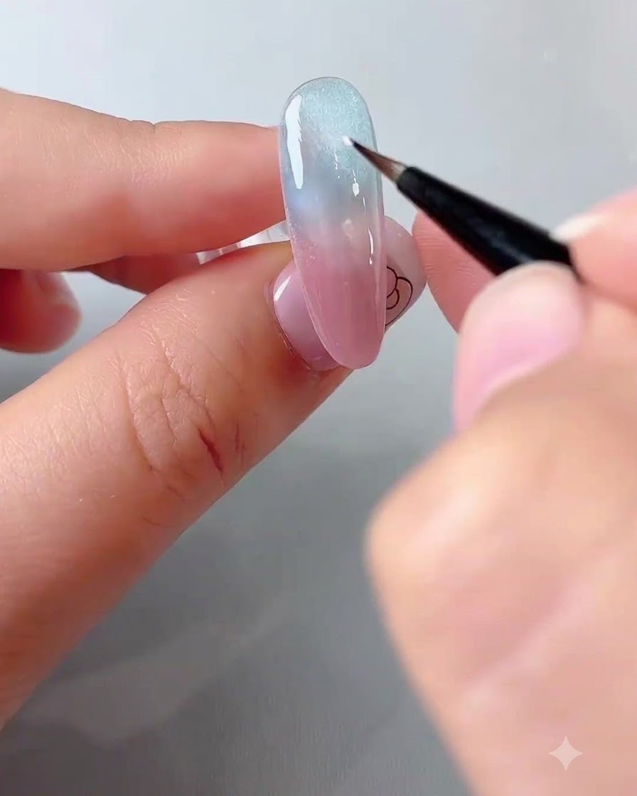 Eine Nail-Künstlerin verwendet einen feinen Pinsel, um einen Klecks weißes Gel auf einen blau-rosa Farbverlaufsnagel zu setzen.